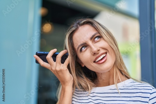 Young blonde woman miling confident listening audio message by the smartphone at street