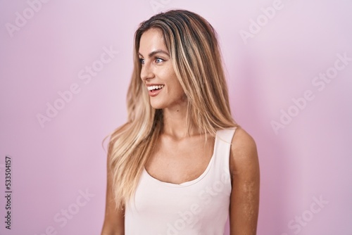 Young blonde woman standing over pink background looking away to side with smile on face, natural expression. laughing confident.