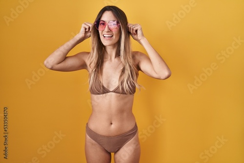 Young hispanic woman wearing bikini over yellow background smiling pulling ears with fingers, funny gesture. audition problem