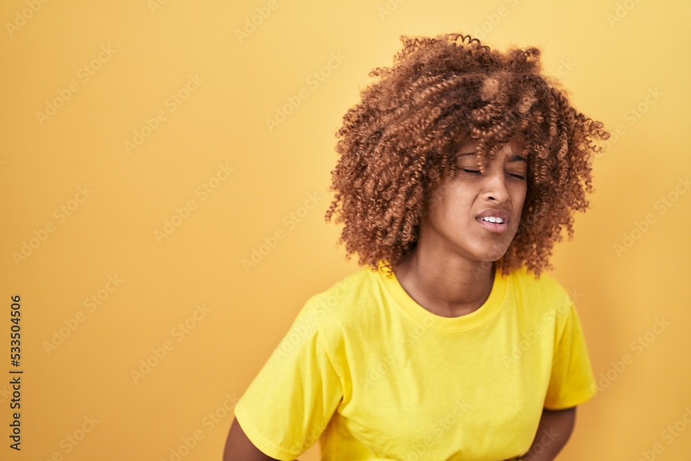 Young hispanic woman with curly hair standing over yellow background with hand on stomach because indigestion, painful illness feeling unwell. ache concept. Young hispanic woman with curly hair standing over yellow background with hand on stomach because indigestion, painful illness feeling unwell. ache concept.