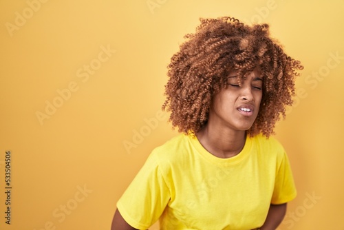 Young hispanic woman with curly hair standing over yellow background with hand on stomach because indigestion, painful illness feeling unwell. ache concept.