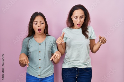 Young mother and daughter standing over pink background pointing down with fingers showing advertisement, surprised face and open mouth