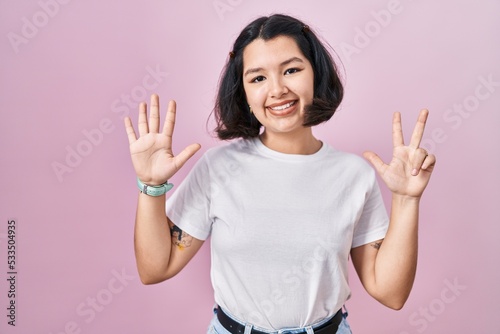 Young hispanic woman wearing casual white t shirt over pink background showing and pointing up with fingers number eight while smiling confident and happy. © Krakenimages.com