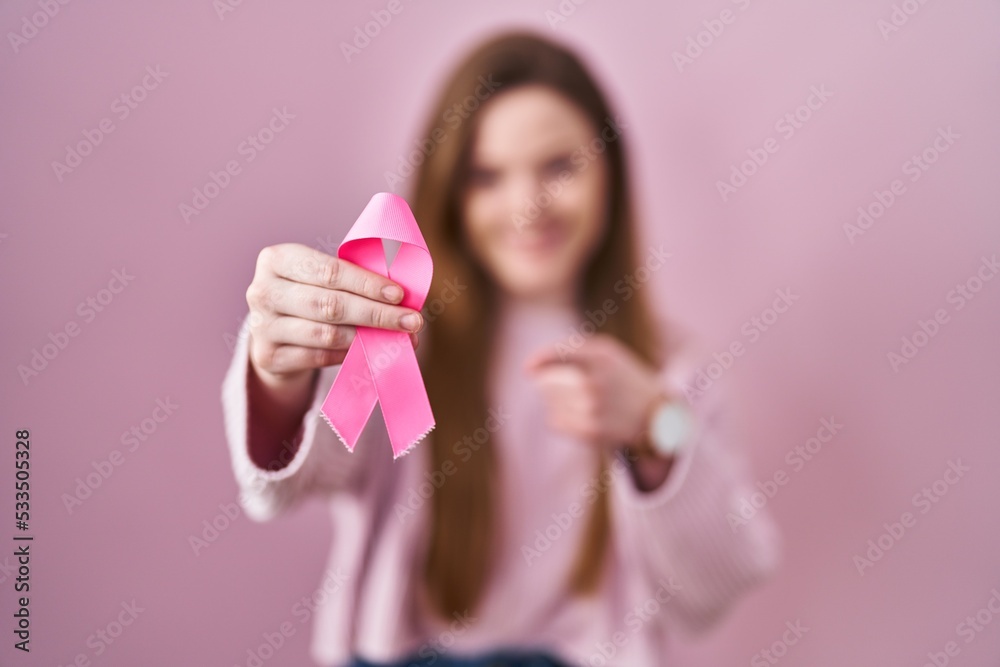 Young caucasian woman holding pink cancer ribbon smiling happy pointing with hand and finger Young caucasian woman holding pink cancer ribbon smiling happy pointing with hand and finger