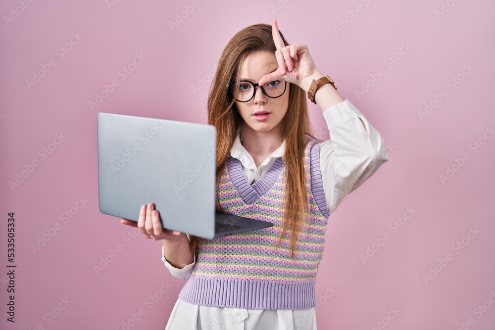 Young caucasian woman working using computer laptop making fun of people with fingers on forehead doing loser gesture mocking and insulting. Young caucasian woman working using computer laptop making fun of people with fingers on forehead doing loser gesture mocking and insulting.