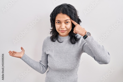 Hispanic woman with dark hair standing over isolated background confused and annoyed with open palm showing copy space and pointing finger to forehead. think about it.