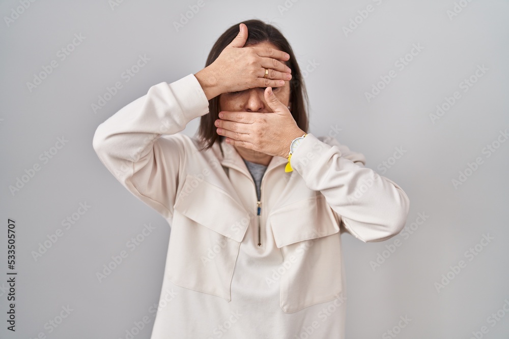 Middle age hispanic woman standing over isolated background covering eyes and mouth with hands, surprised and shocked. hiding emotion Middle age hispanic woman standing over isolated background covering eyes and mouth with hands, surprised and shocked. hiding emotion