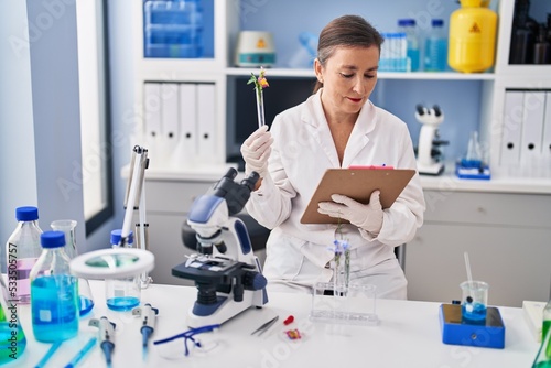 Middle age woman scientist holding test tube with flower reading clipboard at laboratory
