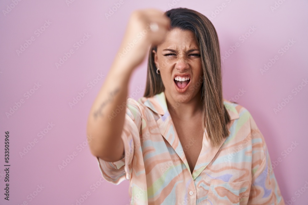 Blonde woman standing over pink background angry and mad raising fist frustrated and furious while shouting with anger. rage and aggressive concept. Blonde woman standing over pink background angry and mad raising fist frustrated and furious while shouting with anger. rage and aggressive concept.