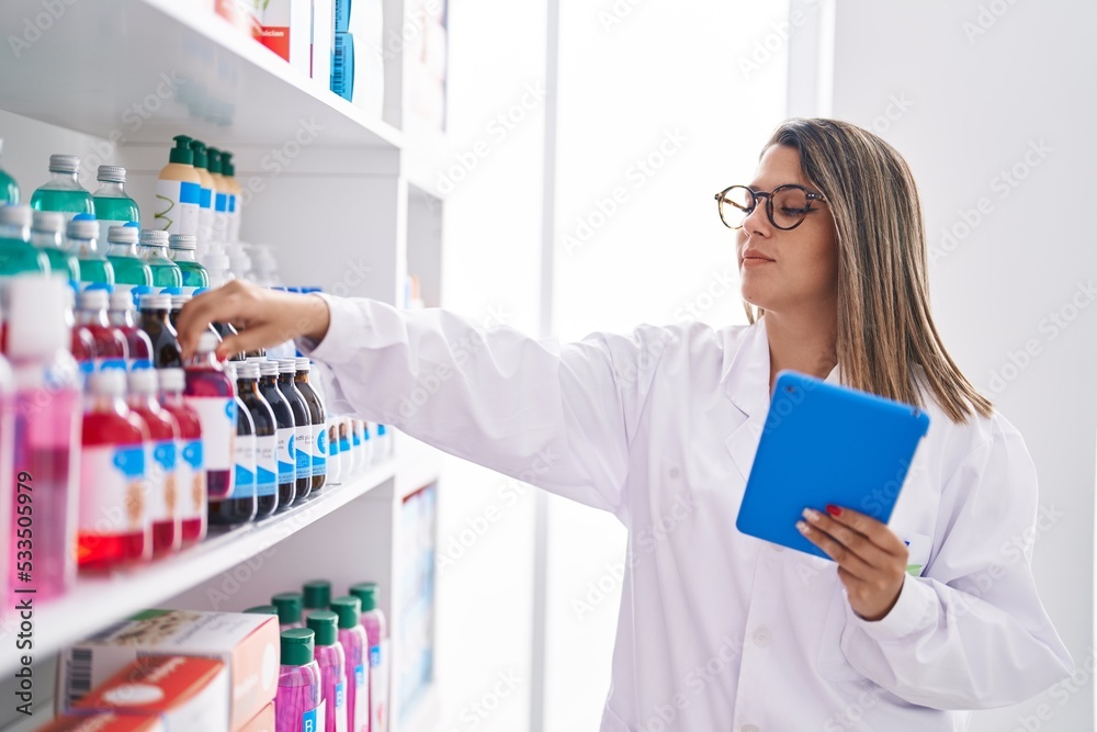Young hispanic woman pharmacist using touchpad holding medicine bottle at pharmacy