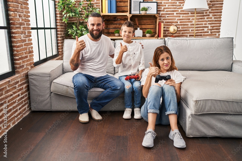 Family of three playing video game sitting on the sofa smiling happy and positive, thumb up doing excellent and approval sign
