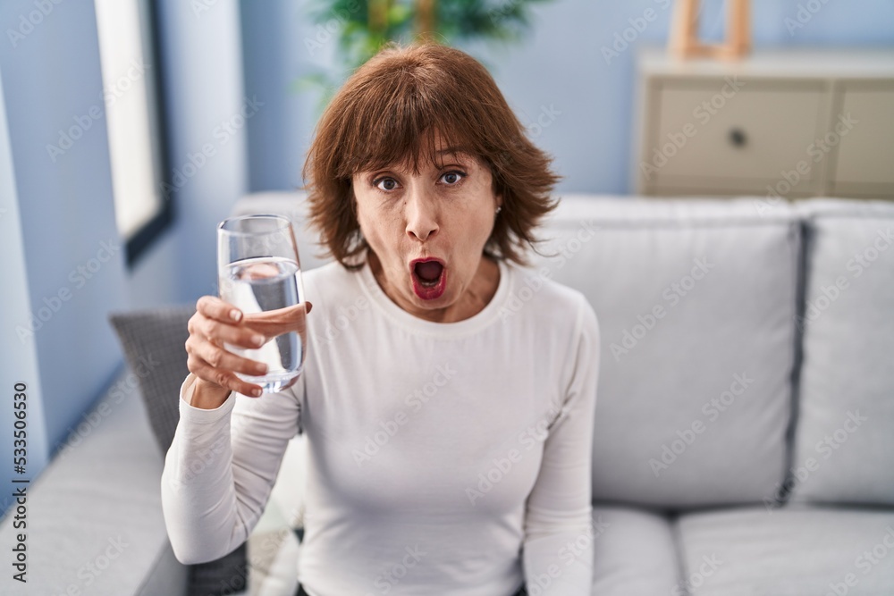 Middle age woman drinking glass of water scared and amazed with open mouth for surprise, disbelief face