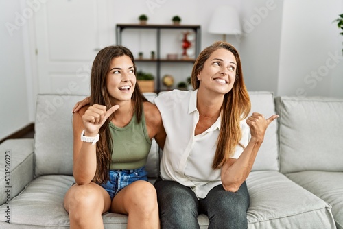 Mother and daughter together sitting on the sofa at home smiling with happy face looking and pointing to the side with thumb up.