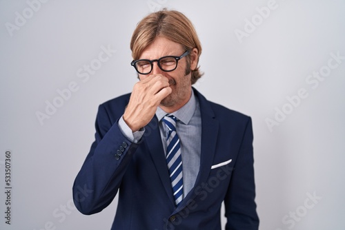 Caucasian man with mustache wearing business clothes smelling something stinky and disgusting, intolerable smell, holding breath with fingers on nose. bad smell
