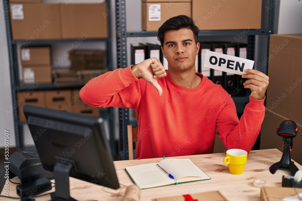 Hispanic man working at small business ecommerce holding open banner with angry face, negative sign showing dislike with thumbs down, rejection concept Hispanic man working at small business ecommerce holding open banner with angry face, negative sign showing dislike with thumbs down, rejection concept