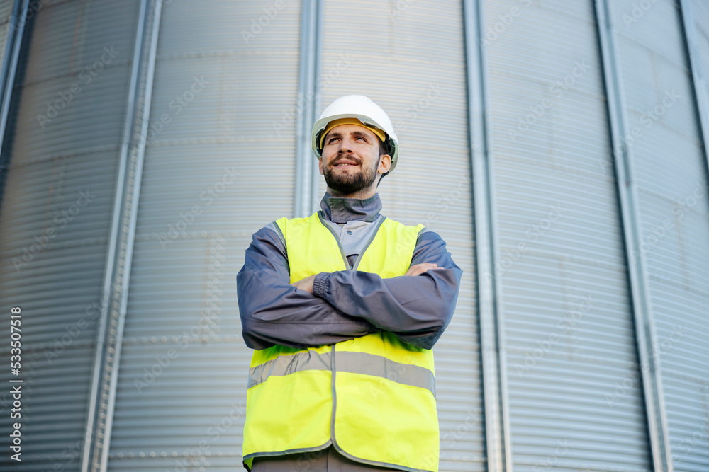 A refinery worker stands outside with arms crossed and smiles in front of the silo.