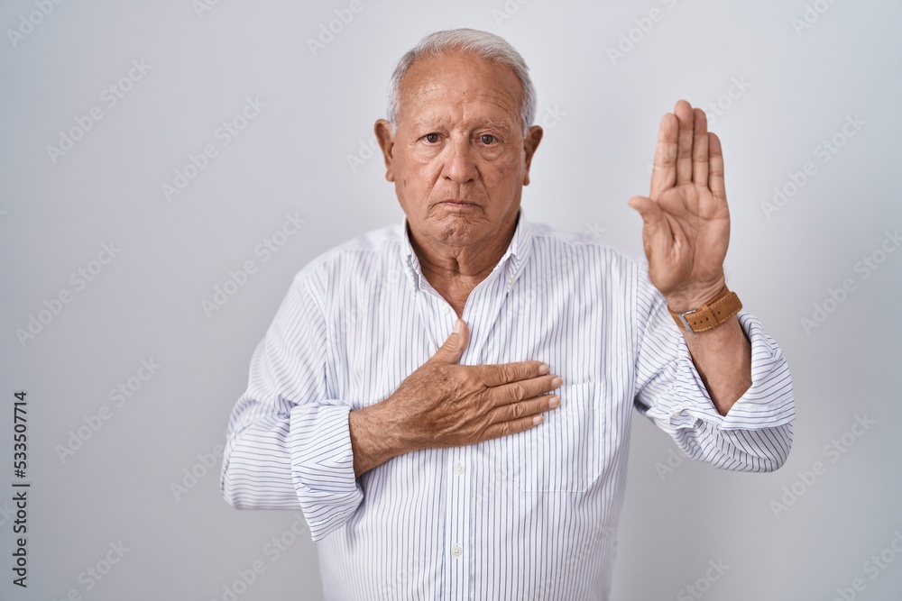 Senior man with grey hair standing over isolated background swearing with hand on chest and open palm, making a loyalty promise oath Senior man with grey hair standing over isolated background swearing with hand on chest and open palm, making a loyalty promise oath