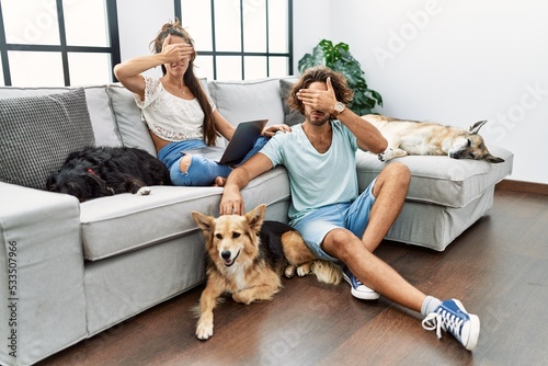 Young hispanic couple with dogs relaxing at home covering eyes with hand, looking serious and sad. sightless, hiding and rejection concept
