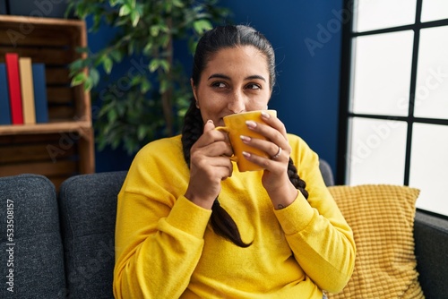 Young hispanic woman drinking coffee sitting on sofa at home