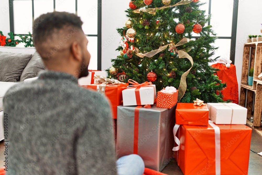 Young african american man on back view looking christmas tree sitting on the floor at home. Young african american man on back view looking christmas tree sitting on the floor at home.