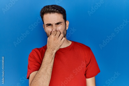 Young hispanic man with beard wearing red t shirt over blue background smelling something stinky and disgusting, intolerable smell, holding breath with fingers on nose. bad smell