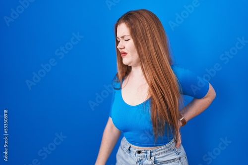 Redhead woman standing over blue background suffering of backache, touching back with hand, muscular pain