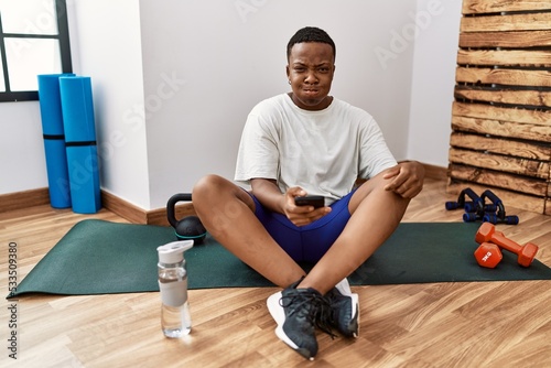 Young african man sitting on training mat at the gym using smartphone puffing cheeks with funny face. mouth inflated with air, crazy expression.