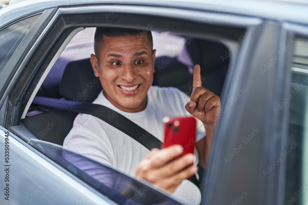 Young hispanic man doing video call with smartphone in the car smiling with an idea or question pointing finger with happy face, number one