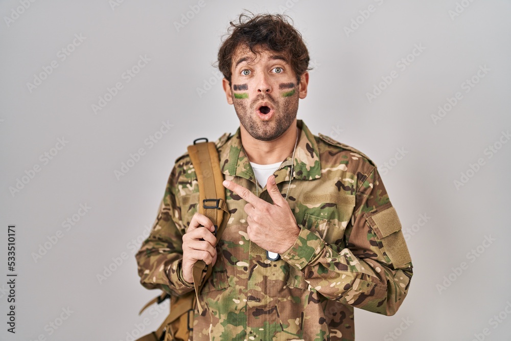 Hispanic young man wearing camouflage army uniform surprised pointing with finger to the side, open mouth amazed expression.