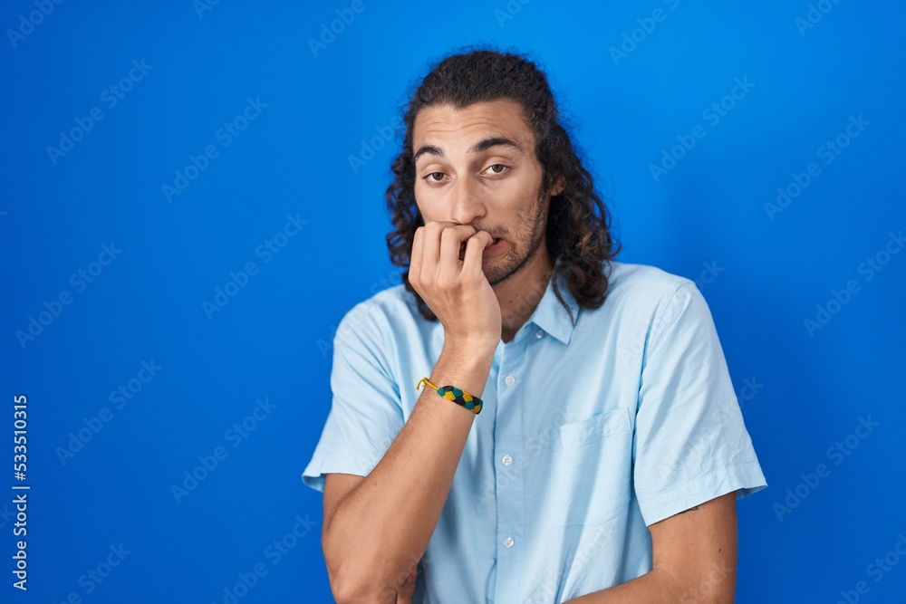 Young hispanic man standing over blue background looking stressed and nervous with hands on mouth biting nails. anxiety problem.