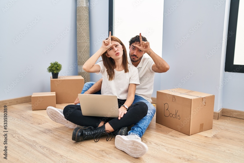 Young couple using laptop at new home making fun of people with fingers on forehead doing loser gesture mocking and insulting. Young couple using laptop at new home making fun of people with fingers on forehead doing loser gesture mocking and insulting.