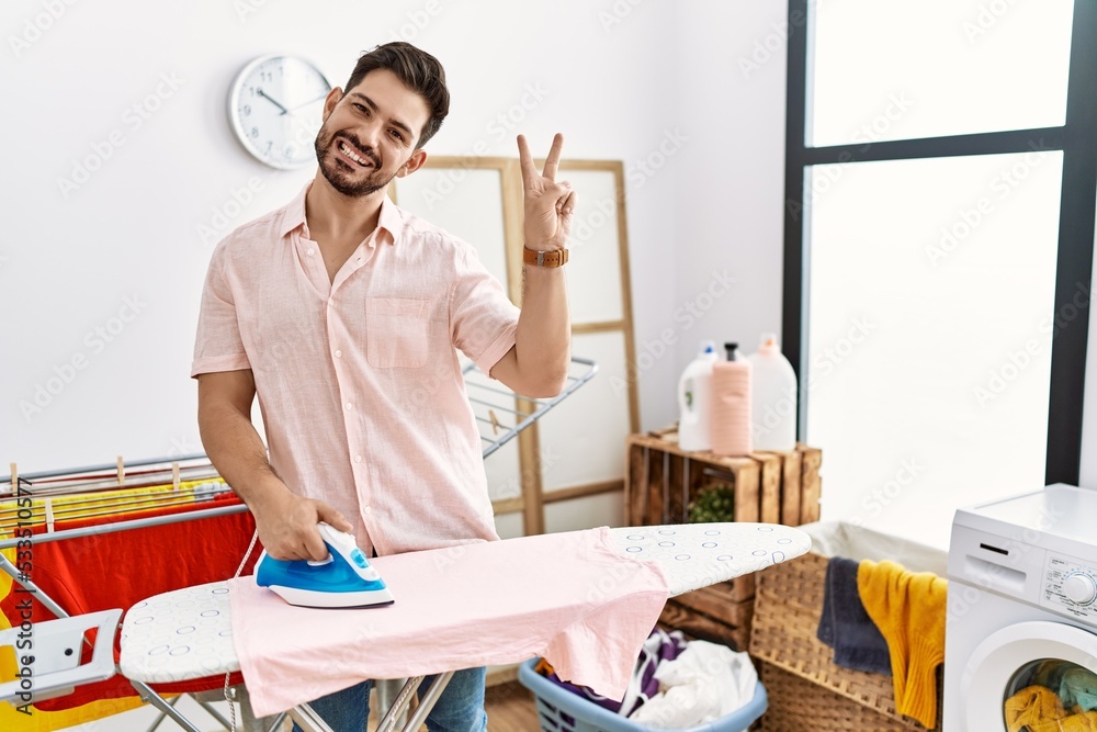 Young man with beard ironing clothes at home smiling looking to the camera showing fingers doing victory sign. number two. Young man with beard ironing clothes at home smiling looking to the camera showing fingers doing victory sign. number two.