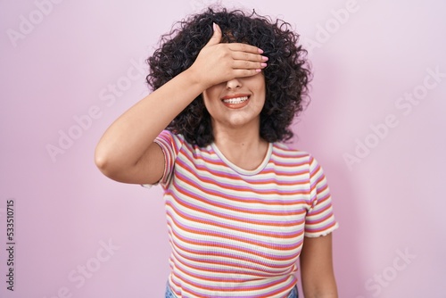 Young middle east woman standing over pink background smiling and laughing with hand on face covering eyes for surprise. blind concept.