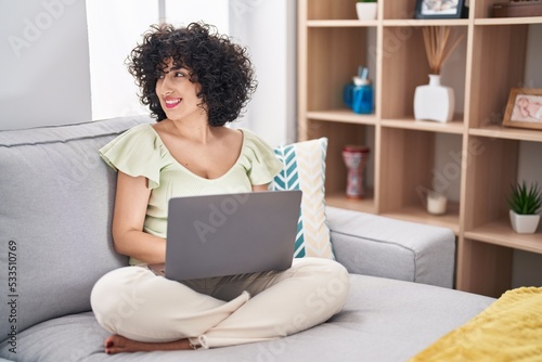 Young brunette woman with curly hair using laptop sitting on the sofa at home looking away to side with smile on face, natural expression. laughing confident.