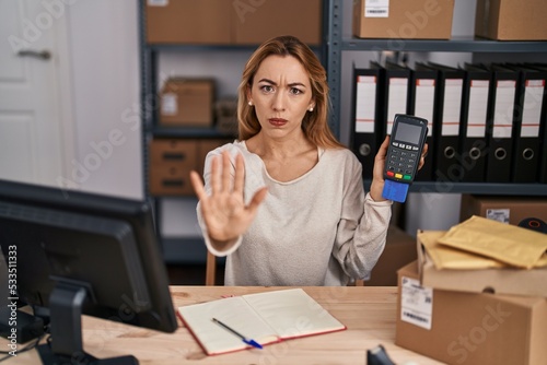 Hispanic woman working at small business ecommerce holding credit card and dataphone with open hand doing stop sign with serious and confident expression, defense gesture