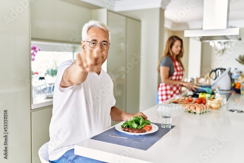 Middle age hispanic couple eating healthy meal at home pointing with finger to the camera and to you, confident gesture looking serious