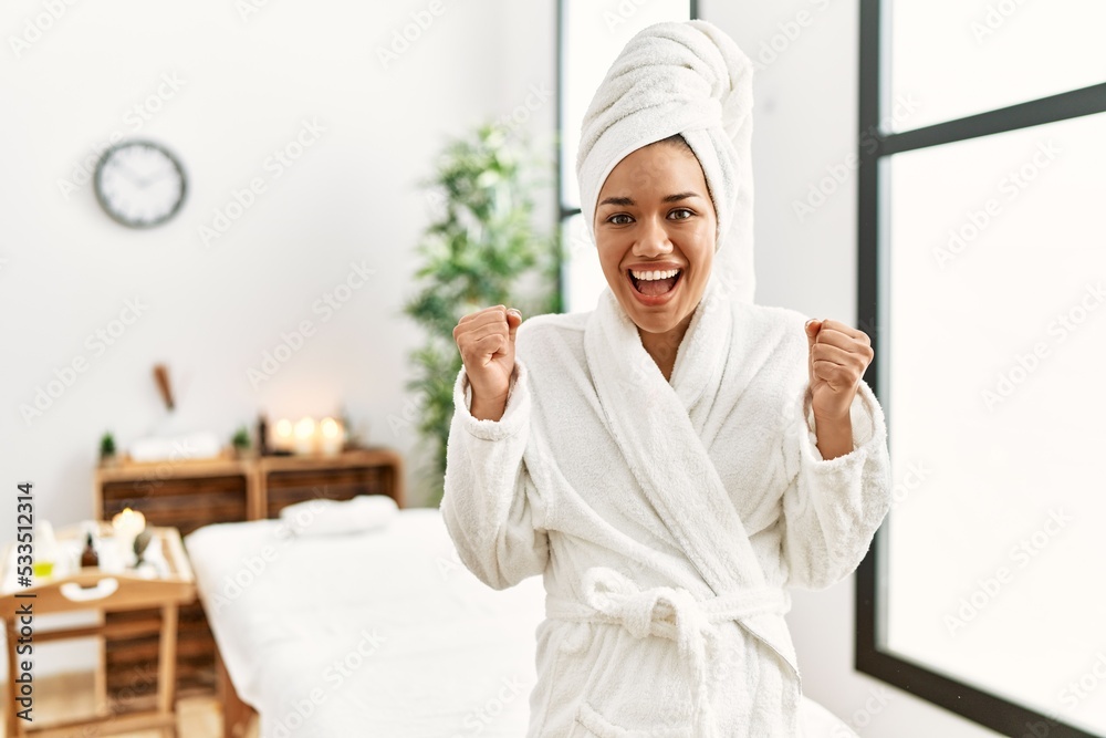 Young brunette woman wearing towel and bathrobe standing at beauty center celebrating surprised and amazed for success with arms raised and open eyes. winner concept.