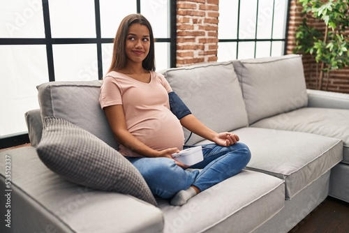Young pregnant woman using blood pressure monitor sitting on the sofa smiling looking to the side and staring away thinking.
