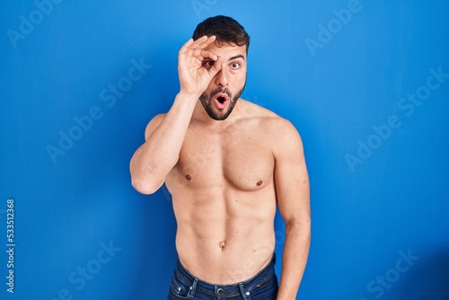 Handsome hispanic man standing shirtless doing ok gesture shocked with surprised face, eye looking through fingers. unbelieving expression.