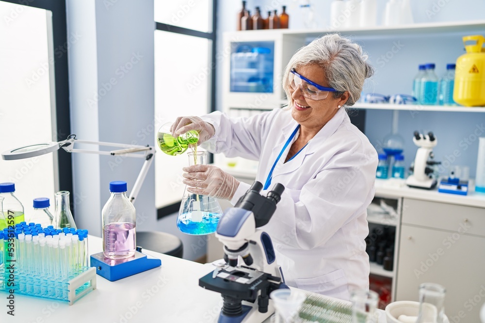 Middle age woman wearing scientist uniform pouring liquid on test tube at laboratory Middle age woman wearing scientist uniform pouring liquid on test tube at laboratory