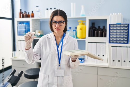 Young hispanic woman working at scientist laboratory with blood samples clueless and confused expression. doubt concept.