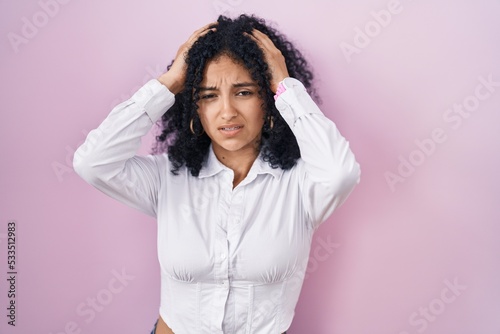 Hispanic woman with curly hair standing over pink background suffering from headache desperate and stressed because pain and migraine. hands on head.