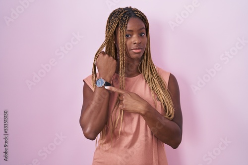 African american woman with braided hair standing over pink background in hurry pointing to watch time, impatience, looking at the camera with relaxed expression