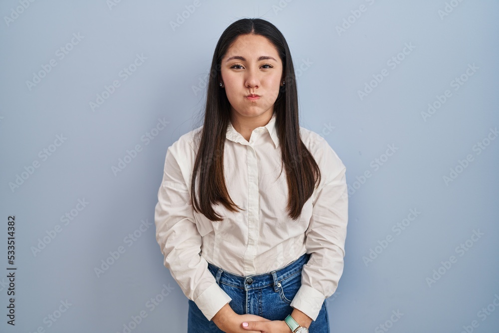Young latin woman standing over blue background puffing cheeks with funny face. mouth inflated with air, crazy expression. Young latin woman standing over blue background puffing cheeks with funny face. mouth inflated with air, crazy expression.