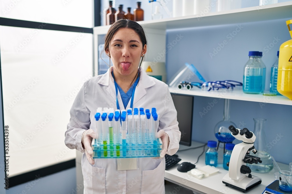 Young latin woman working at scientist laboratory sticking tongue out happy with funny expression.