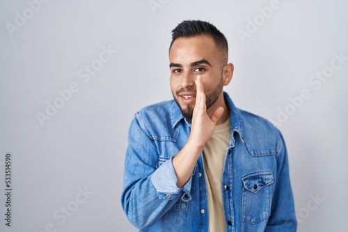 Young hispanic man standing over isolated background hand on mouth telling secret rumor, whispering malicious talk conversation