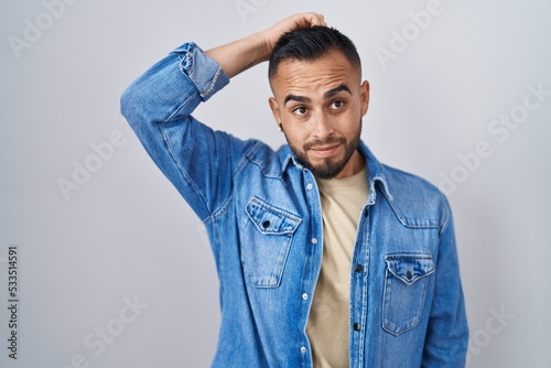 Young hispanic man standing over isolated background confuse and wondering about question. uncertain with doubt, thinking with hand on head. pensive concept.