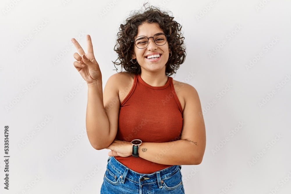 Young hispanic woman wearing glasses standing over isolated background smiling with happy face winking at the camera doing victory sign with fingers. number two. Young hispanic woman wearing glasses standing over isolated background smiling with happy face winking at the camera doing victory sign with fingers. number two.