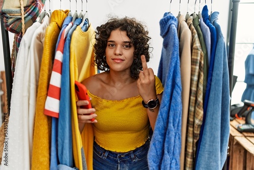 Young hispanic woman searching clothes on clothing rack using smartphone showing middle finger, impolite and rude fuck off expression