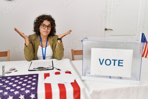 Young hispanic woman at political election sitting by ballot clueless and confused expression with arms and hands raised. doubt concept. photo